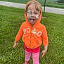 child, orange_hoodie, pink_leggings, grass, outdoor, face_paint, tongue_out, playful, smiling, road, trees, field, casual_clothing, footwear, person, happy, nature, fun, expression, summer