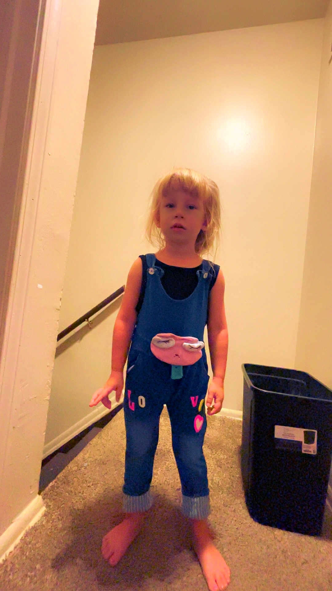 Alaiya Mary Deeter joined the competition — help win amazing prizes! child, barefoot, overalls, carpet, hallway, wall, stairs, railing, trash_bin, blue_clothing, standing, indoor, young_child, person, feet, casual, home, light_skin, blonde_hair, expressionless