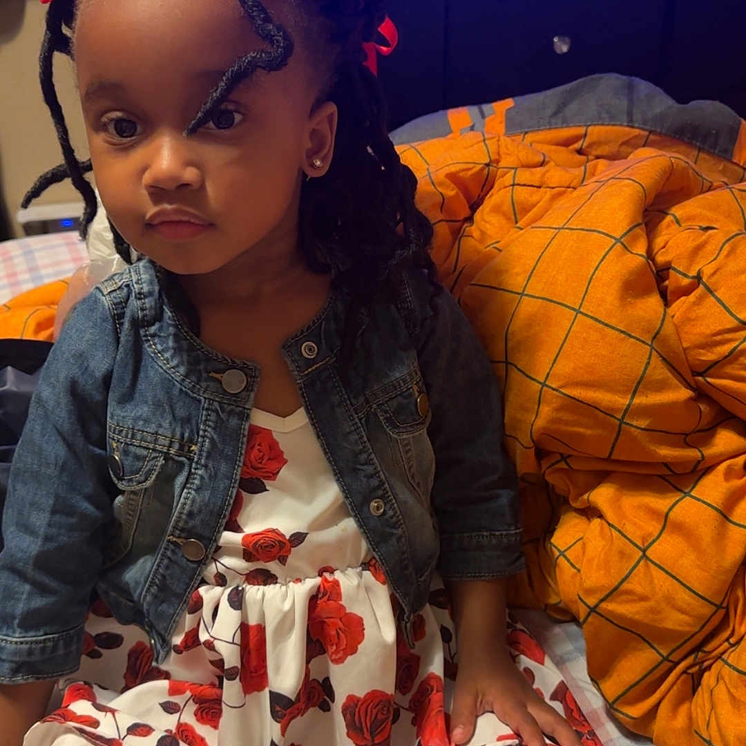 Morgan joined the competition — help win amazing prizes! bed, blanket, braided_hair, casual, child, clothing, cross_legged, cute, denim_jacket, expression, floral_dress, footwear, girl, home, indoor, orange_bows, portrait, sitting, television, young_child