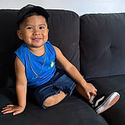 Eiden joined the competition — help win amazing prizes! child, smiling, sitting, couch, blue_shirt, denim_shorts, cap, necklace, shoe, indoors, happy, portrait, casual, young_child, black_couch, comfortable, cute, fashion, relaxed, person