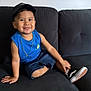 child, smiling, sitting, couch, blue_shirt, denim_shorts, cap, necklace, shoe, indoors, happy, portrait, casual, young_child, black_couch, comfortable, cute, fashion, relaxed, person