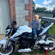 Naël participe au concours pour gagner de l'argent avec cette photo : child, motorcycle, outdoor, daylight, grass, gravel, brick_wall, stone_wall, trailer, blue_sky, clouds, jeans, striped_shirt, footwear, vehicle, tire, head, person, curious, adventure