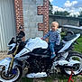 child, motorcycle, outdoor, daylight, grass, gravel, brick_wall, stone_wall, trailer, blue_sky, clouds, jeans, striped_shirt, footwear, vehicle, tire, head, person, curious, adventure