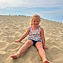 child, girl, sand, dune, barefoot, summer_clothing, floral_top, denim_shorts, outdoor, nature, sky, clouds, happy, smiling, casual, young, portrait, sitting, sunlight, playful