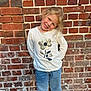 child, girl, blonde_hair, smiling, white_shirt, flower_design, jeans, brick_wall, outdoor, playful_pose, casual_clothing, happy, portrait, daylight, young, person, fashion, cute, standing, head_tilt
