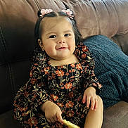 Nahomy joined the competition — help win amazing prizes! toddler, child, smiling, sitting, couch, blanket, cookie, hair_bows, floral_dress, indoor, happy, cute, person, portrait, cozy, furniture, socks, brown, knitted, snack