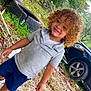 car, casual_clothing, child, curly_hair, daylight, dirt, grass, grey_shirt, happy, leaves, nature, navy_shorts, outdoor, person, playful, smiling, tires, toddler, trees, young_child