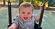 Nikolai is registered to the contest to win money with this photo: child, toddler, swing, playground, outdoor, sunlight, happy, smiling, curly_hair, greenery, park, clothing, short_sleeves, metal_chain, seat, nature, daylight, fence, bench, shadow