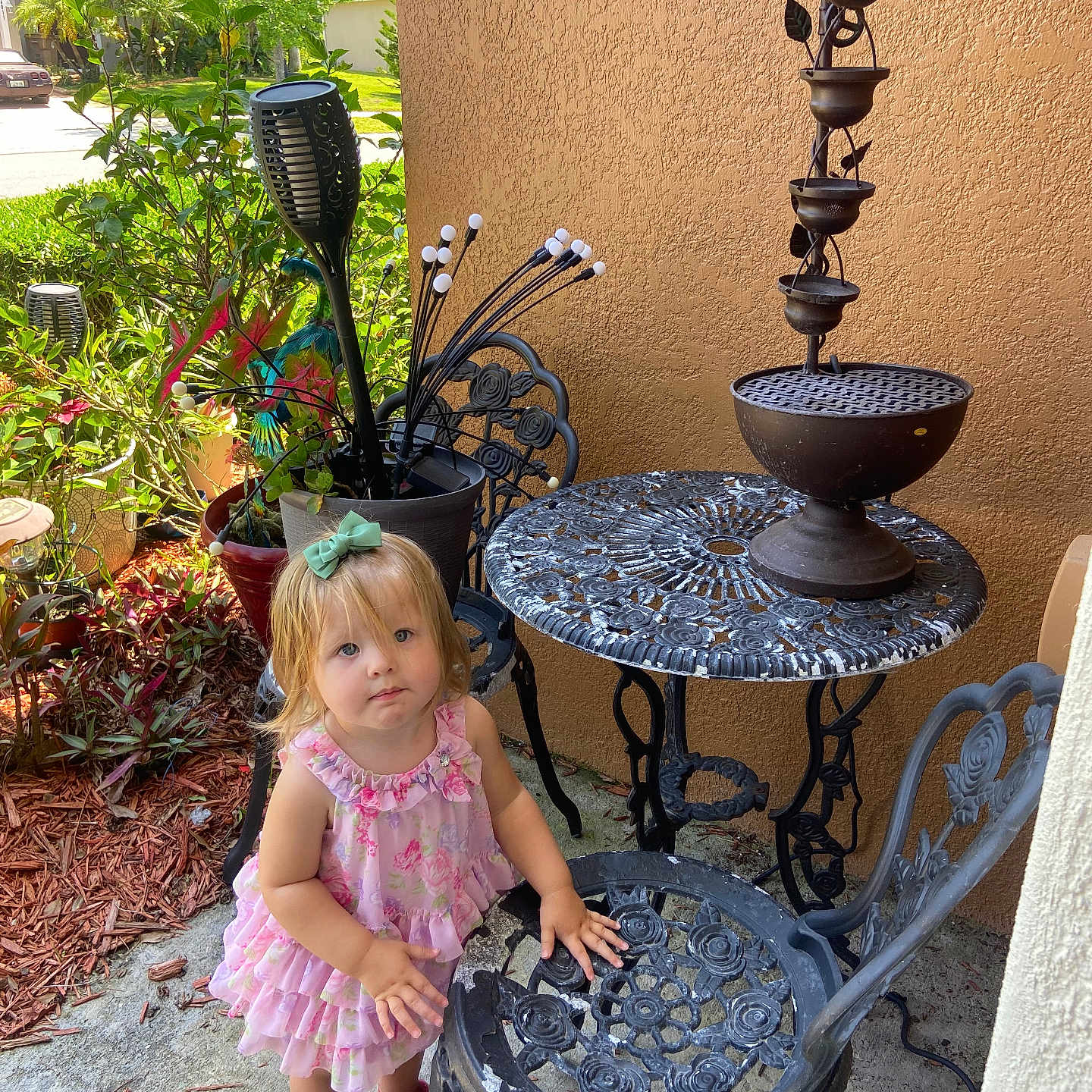 Nalla is registered to the contest to win money with this photo: blond_hair, bow_hair_accessory, child, concrete_floor, curious_expression, daylight, flower_pot, garden, greenery, outdoor, patio, pink_dress, pink_shoes, plants, summer, sunlight, toddler, wrought_iron_chair, wrought_iron_table, young_child
