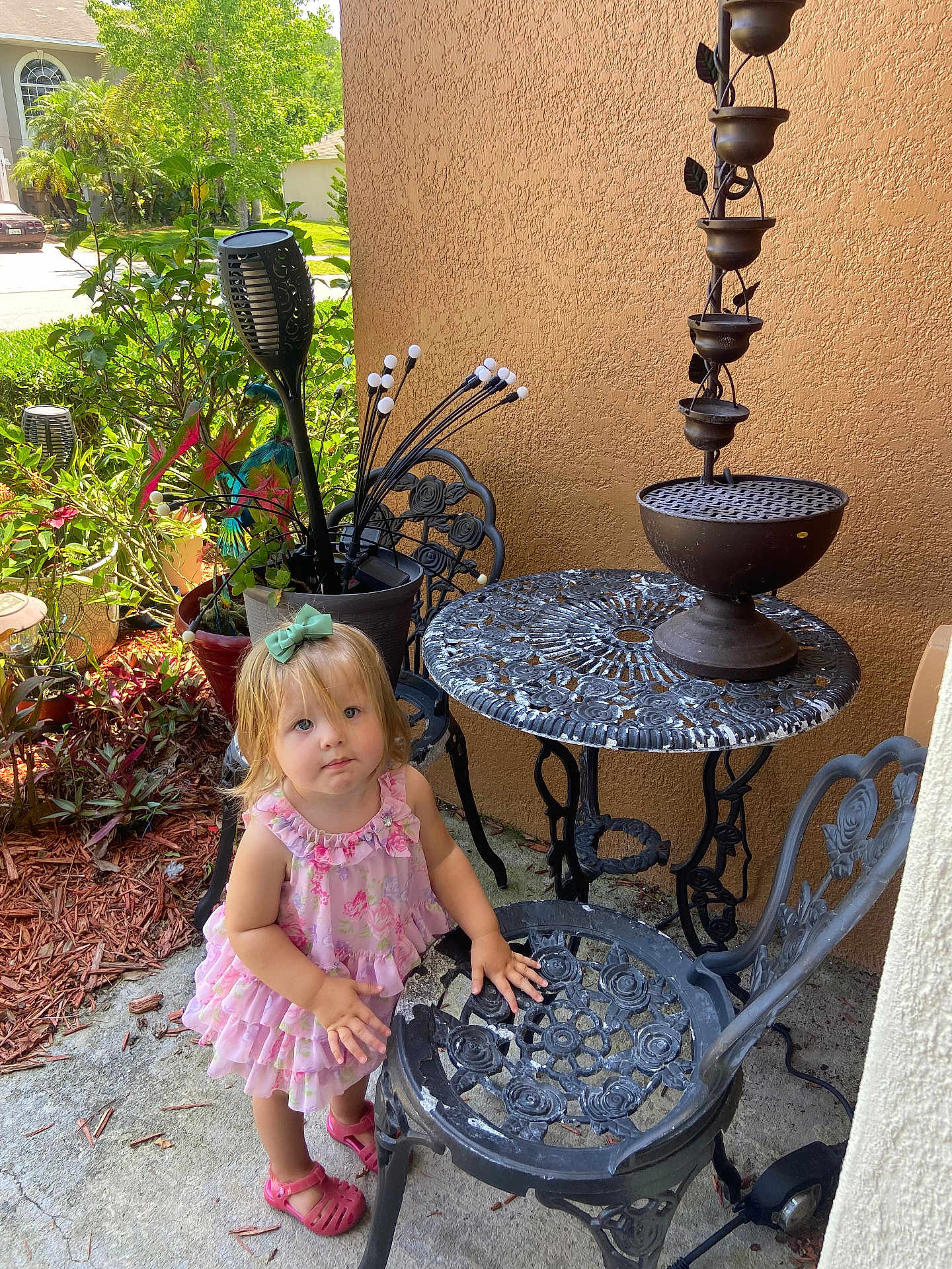 Nalla is registered to the contest to win money with this photo: blond_hair, bow_hair_accessory, child, concrete_floor, curious_expression, daylight, flower_pot, garden, greenery, outdoor, patio, pink_dress, pink_shoes, plants, summer, sunlight, toddler, wrought_iron_chair, wrought_iron_table, young_child