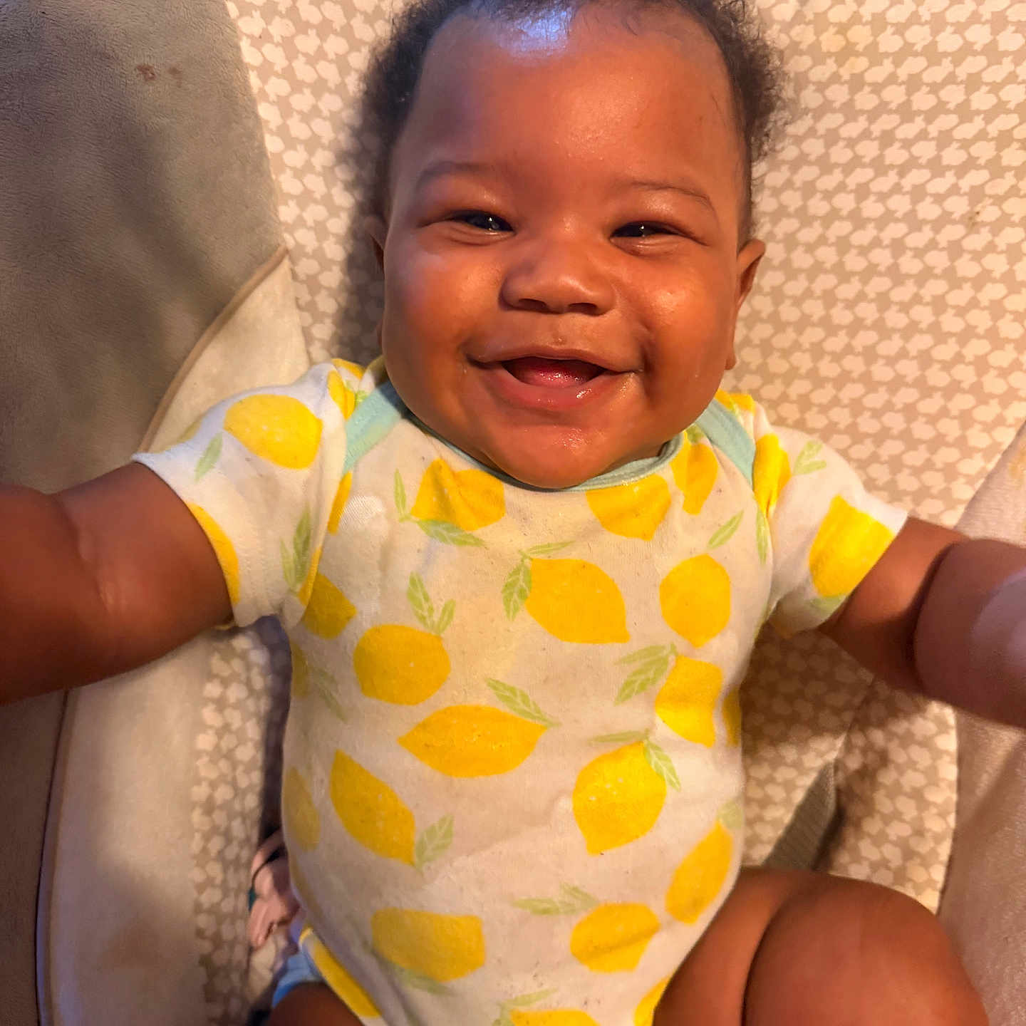 Genesis is registered to the contest to win money with this photo: baby, infant, child, smiling, happy, portrait, closeup, face, sitting, indoors, onesie, lemon_pattern, yellow, cushion, blanket, chair, curly_hair, dark_skin, adorable, photography