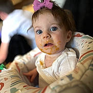 Kinzley is registered to the contest to win money with this photo: baby, child, high_chair, food, messy_face, purple_bow, blue_eyes, white_clothing, indoor, person, infant, curious, cute, mess, feeding_time, soft_focus, portrait, young_child, seated, colorful