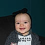 baby, black_bow, cardigan, child, closeup, couch, cute, dark_background, face, flower_pattern, happy, headband, indoor, infant, portrait, seated, smiling, teal_wall, white_shirt, young_child
