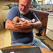 Alana joined the competition — help win amazing prizes! man, baby, holding, kitchen, wooden_cabinets, sink, fan, remote_control, table, jeans, tshirt, watch, floor, window, dish_rack, bottle, tattoo, facial_hair, indoor, person