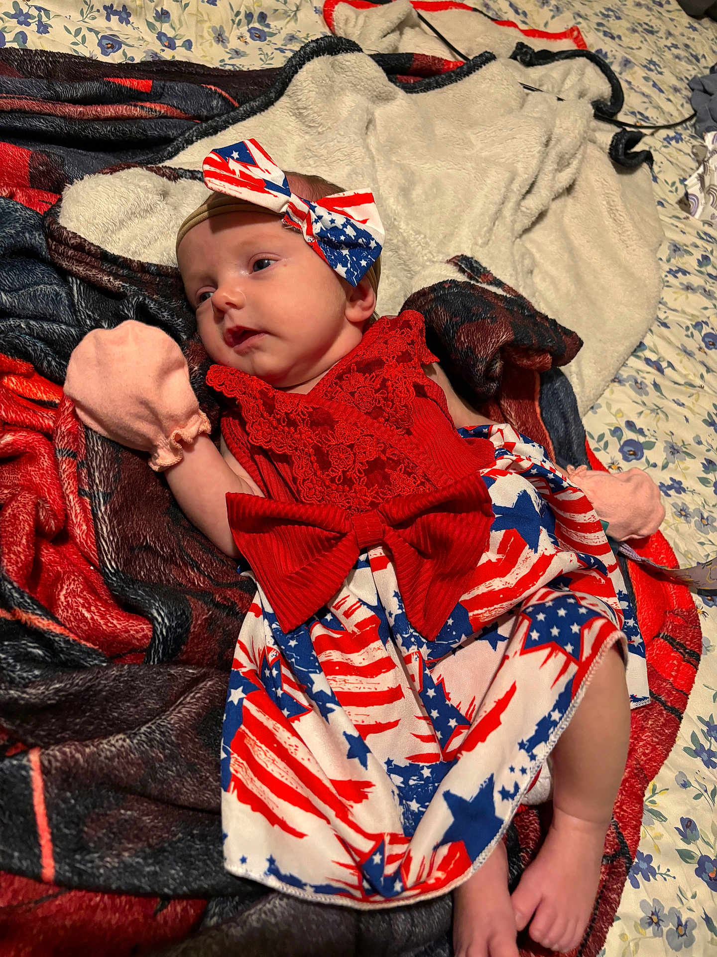 Alana is registered to the contest to win money with this photo: baby, infant, red_dress, headband, bow, mittens, blanket, bed, floral_sheet, foot, lying_down, cute, patterned_clothing, stars, stripes, sleepwear, soft_texture, indoors, newborn, portrait