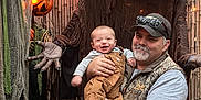 Wesley is registered to the contest to win money with this photo: baby, man, pumpkin, halloween, costume, scary, outdoor, decoration, smiling, face, child, adult, festive, orange, lights, wooden_fence, vest, socks, cap, overalls