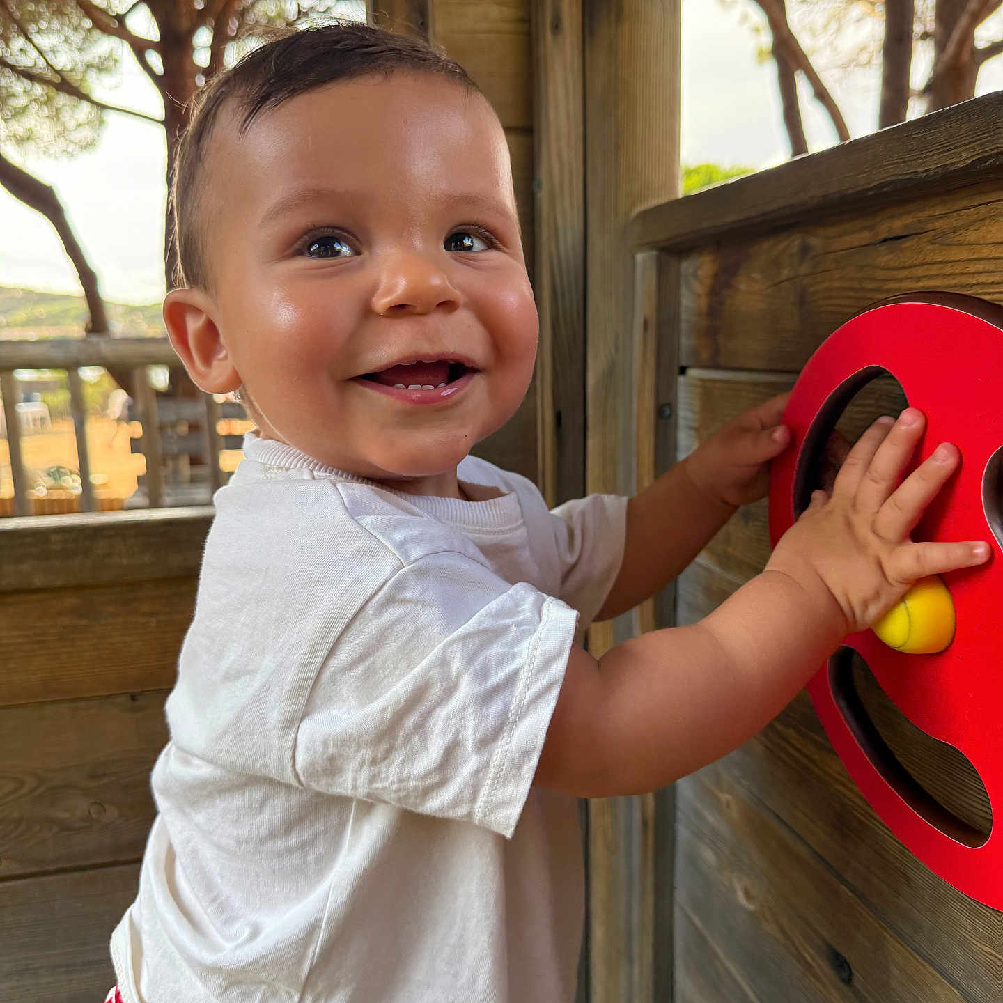 Marceau a rejoint le concours — aidez-le/la à gagner de superbes lots ! casual_clothing, child, closeup, daylight, face, hands, happy, nature, outdoor, person, playground, playing, red_striped_shorts, red_wheel_toy, short_hair, smiling, toddler, trees, white_shirt, wooden_play_structure