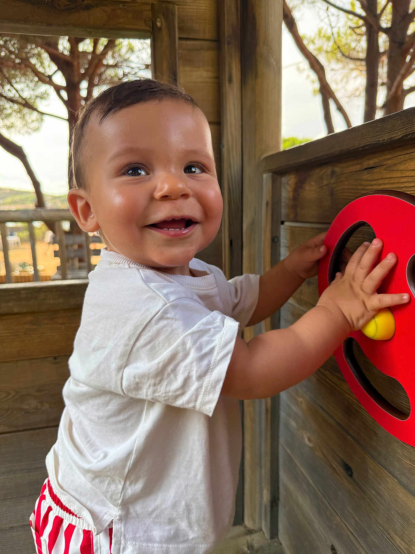 Marceau a rejoint le concours — aidez-le/la à gagner de superbes lots ! toddler, child, smiling, white_shirt, red_striped_shorts, wooden_play_structure, red_wheel_toy, outdoor, playground, happy, hands, face, short_hair, daylight, nature, trees, person, playing, closeup, casual_clothing