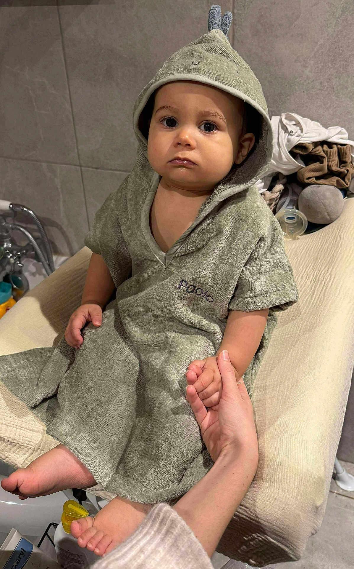 Paolo participe au concours pour gagner de l'argent avec cette photo : baby, infant, toddler, hooded_towel, towel, bath_time, changing_table, tile_wall, holding_hand, adult_hand, bare_feet, pacifier, plush_toy, clothing, bathroom, portrait, serious_expression, big_eyes, sitting, soft_lighting