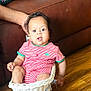 baby, infant, child, striped_clothing, wicker_basket, furniture, brown_couch, wooden_floor, hand, indoor, colorful_pillow, toy_cart, cute, seated, expression, living_room, person, supporting_hand, casual, home