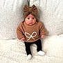 baby, child, knitwear, headband, bow, sweater, black_pants, white_socks, couch, cushion, soft_texture, indoor, portrait, cute, infant, seated, looking_at_camera, cozy, warm_clothing, neutral_colors