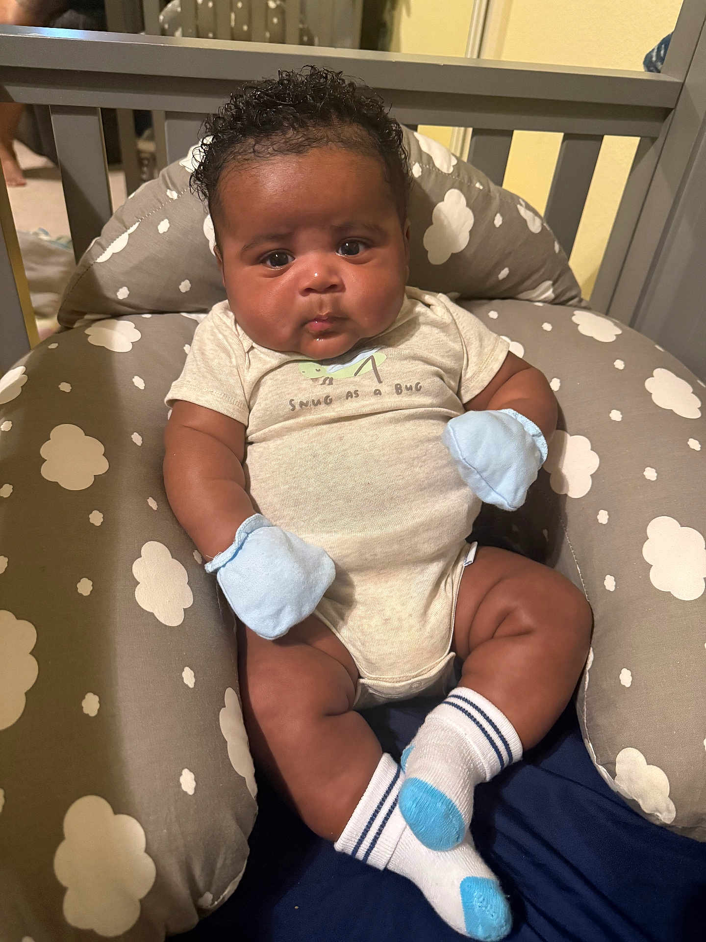 Tyler joined the competition — help win amazing prizes! baby, infant, child, curly_hair, mittens, socks, onesie, cushion, cloud_pattern, gray, beige, indoor, crib, person, cute, plush, seated, looking, portrait, soft_lighting