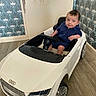 baby, child, toy_car, audi, indoor, flooring, wallpaper, blue_clothing, sitting, steering_wheel, curious_expression, small_wheels, floor, car_toy, person, head, face, seat, vehicle, play