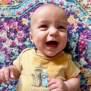 Silas is registered to the contest to win money with this photo: baby, smiling, infant, yellow_shirt, cute, happy, patterned_rug, colorful, lying_down, portrait, child, indoors, clothing, person, face, hand, skin, head, expression, joy