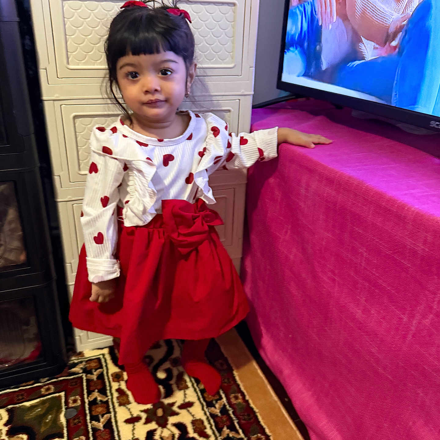 Maryam joined the competition — help win amazing prizes! child, girl, red_skirt, heart_pattern, long_sleeves, standing, indoor, carpet, cabinet, television, pink_table_cover, black_hair, pigtails, ribbons, earrings, expression, home, decor, socks, person