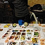 board_game, bottle, cards, cat, chair, cozy, cup, curly_hair, decor, game_pieces, glasses, indoor, paper, person, plastic_chair, playing, relaxed, table, tokens, wooden_furniture