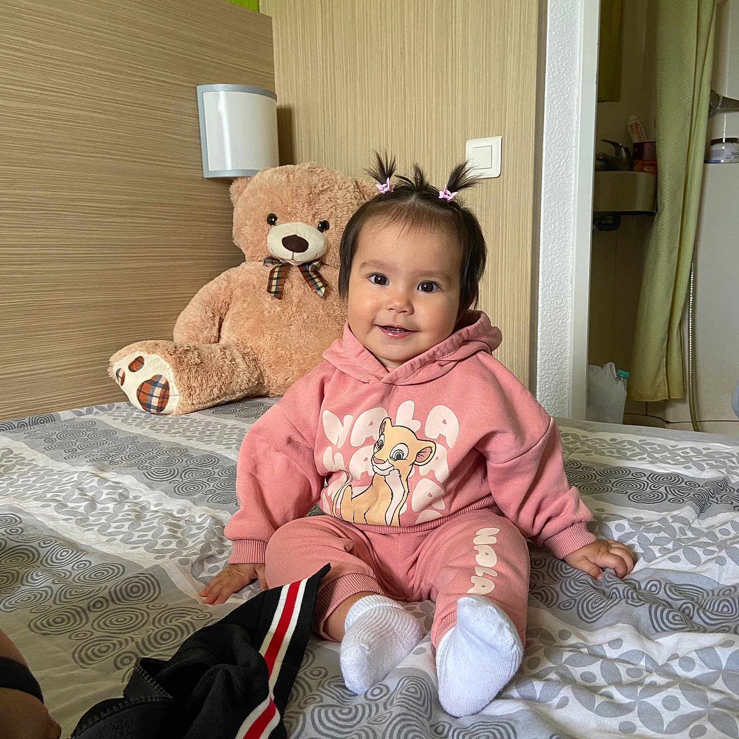Amilia participe au concours pour gagner de l'argent avec cette photo : baby, bed, bedspread, child, curtain, cute, hair_ties, hoodie, indoor, light_switch, pants, patterned_bedspread, person, pink_clothing, room, smiling, teddy_bear, toy, wall, white_socks