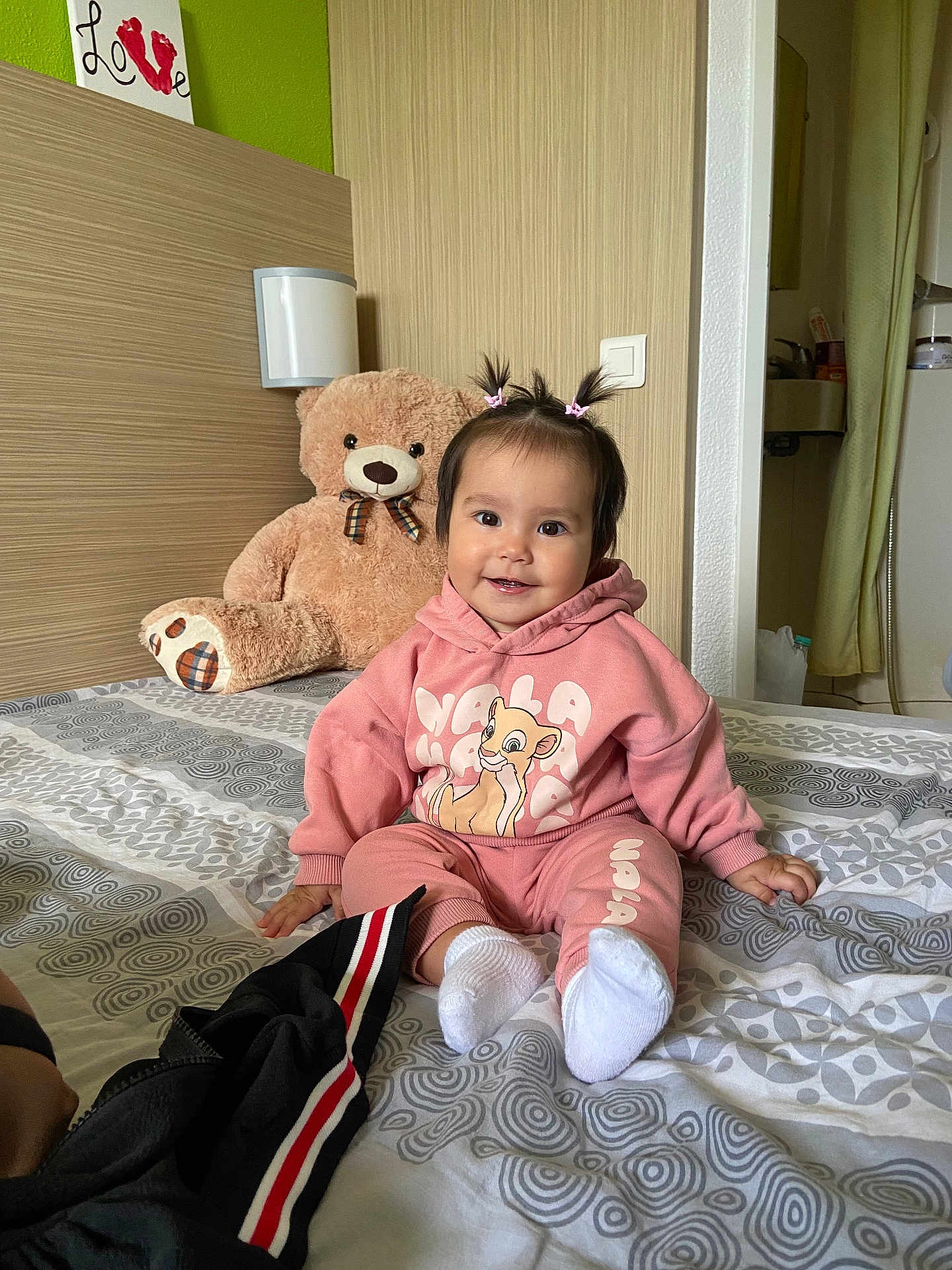 Amilia participe au concours pour gagner de l'argent avec cette photo : baby, bed, bedspread, child, curtain, cute, hair_ties, hoodie, indoor, light_switch, pants, patterned_bedspread, person, pink_clothing, room, smiling, teddy_bear, toy, wall, white_socks