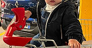 Demir a rejoint le concours — aidez-le/la à gagner de superbes lots ! toddler, child, glasses, shopping_cart, parking_garage, car, concrete, pillar, jacket, denim_overall, sneakers, toy, hand, face, person, urban, indoor, expression, candid, retail