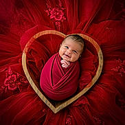 Zaiden is registered to the contest to win money with this photo: baby, infant, swaddled, red_wrap, heart_shaped_bowl, wooden_bowl, smiling, face, cute, newborn, portrait, fabric, red_fabric, floral_embroidery, soft_texture, laying_down, close_up, adorable, happy, child