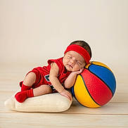 Zaiden is registered to the contest to win money with this photo: baby, infant, sleeping, basketball, red_clothing, headband, pillow, wooden_floor, sports, cute, resting, toddler, child, indoor, soft_texture, colorful, peaceful, small, portrait, cozy
