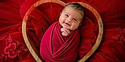 Zaiden joined the competition — help win amazing prizes! baby, infant, swaddled, red_wrap, heart_shaped_bowl, wooden_bowl, smiling, face, cute, newborn, portrait, fabric, red_fabric, floral_embroidery, soft_texture, laying_down, close_up, adorable, happy, child