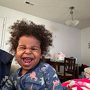 Quincy is registered to the contest to win money with this photo: toddler, child, laughing, curly_hair, indoor, onesie, dinosaur_pattern, blanket, smiling, happy, furniture, table, chair, living_room, person, cozy, casual, home, cute, portrait