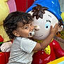 affection, blue_hat, cartoon_character, child, closeup, curly_hair, cute, hugging, indoor, innocence, joy, pacifier, person, playful, red_outfit, smiling, statue, toddler, toy, yellow_scarf