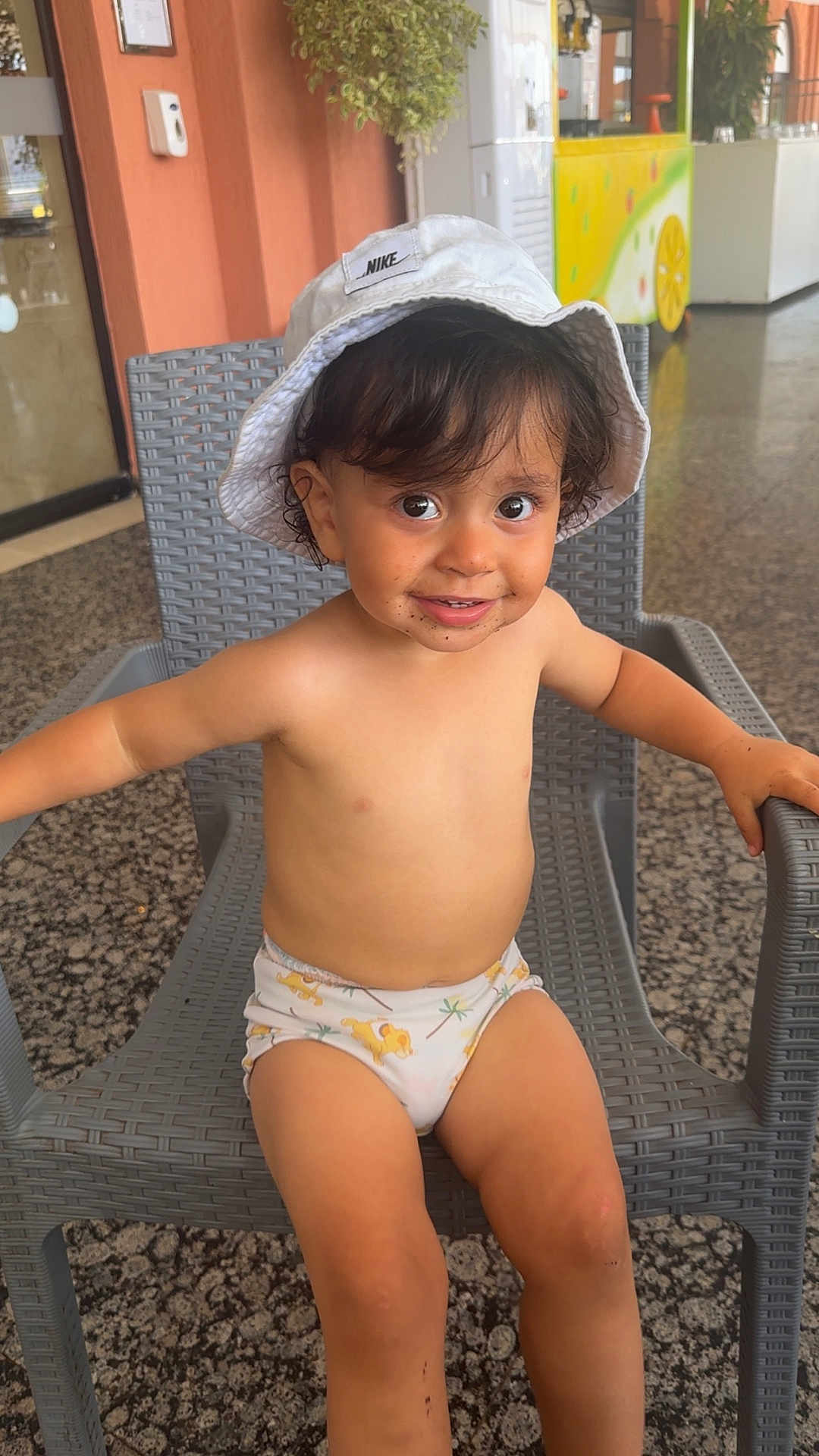 Ayaan participe au concours pour gagner de l'argent avec cette photo : toddler, child, hat, nike, bucket_hat, smile, chair, indoor, swim_diaper, playful, dirt, legs, face, happy, person, wicker_chair, plant, background, colorful, lemon_cart