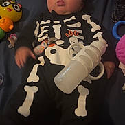 Kaydence is registered to the contest to win money with this photo: baby, infant, child, portrait, lying_down, skeleton_costume, black_outfit, bottle, toys, stuffed_toy, pacifier, smiling, puckered_lips, indoor, blanket, bed, colorful, hearts, emoji, photograph