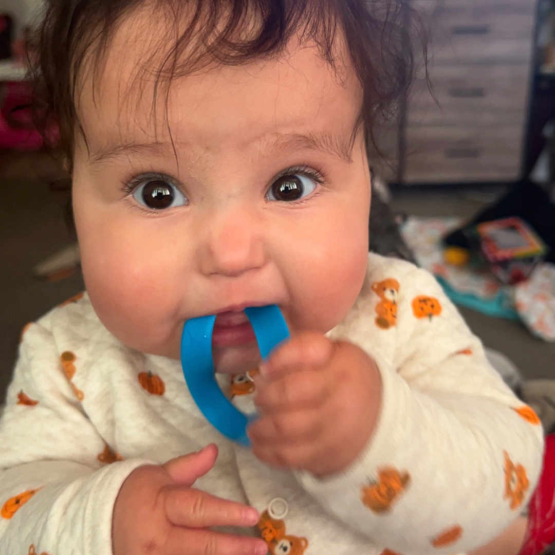 Kaydence is registered to the contest to win money with this photo: baby, infant, child, portrait, close_up, wide_eyed, smiling, sitting, toy, blue_toy, onesie, pumpkin_pattern, clothing, indoor, living_room, television, furniture, home, hands, playful