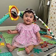 Romina is registered to the contest to win money with this photo: baby, child, girl, purple_dress, floral, headband, daisy, play_mat, mirror_toy, colorful, sitting, smiling, indoor, lamp, curly_hair, cute, happy, toys, floor, portrait