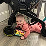 baby, smiling, floor, stroller, wheel, pillow, colorful, pink_clothing, happy, indoors, wood_floor, child, person, crawling, cute, playful, toy, home, young_child, infant