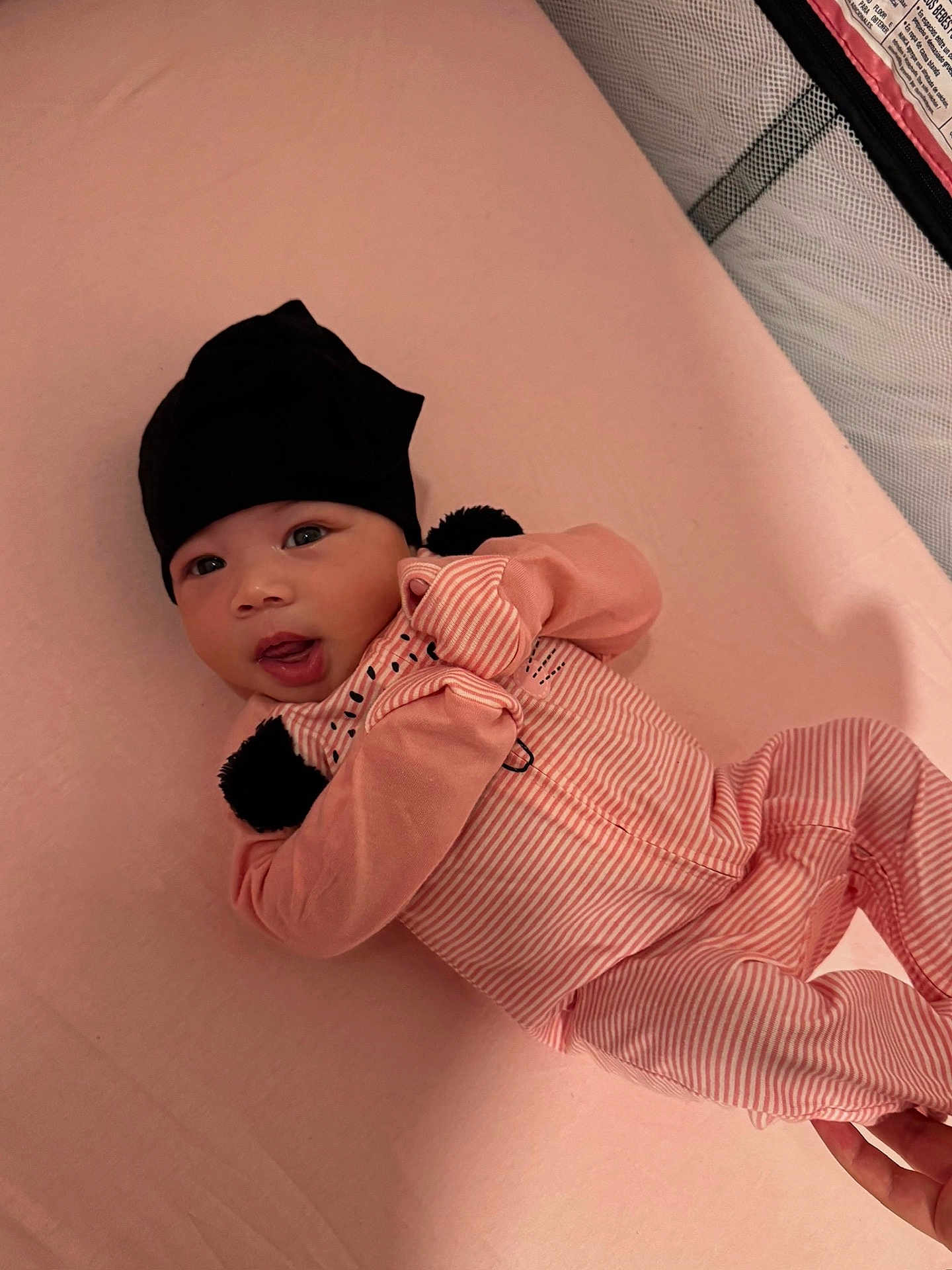 Heaven is registered to the contest to win money with this photo: baby, infant, pink, black_hat, pajamas, striped_clothing, lying_down, soft_surface, cute, child, newborn, indoors, person, face, hand, sleeping_area, cozy, expression, looking_at_camera, comfortable