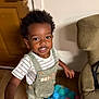toddler, child, smiling, indoor, overalls, striped_shirt, curly_hair, ball_pit, colorful_balls, door, chair, wood_floor, person, happy, young_child, play, home, furniture, casual_clothing, portrait