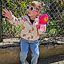 child, sunglasses, bubble_blower, bubbles, jacket, jeans, sneakers, outdoor, sunlight, fence, greenery, play, fun, happy, casual_clothing, flower_pattern, daytime, nature, person, cute