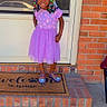 Grace is registered to the contest to win money with this photo: child, girl, smiling, purple_dress, colorful_beads, hair, standing, welcome_mat, brick_wall, porch, door, shoes, outdoor, concrete, happy, person, face, window, casual, sunlight