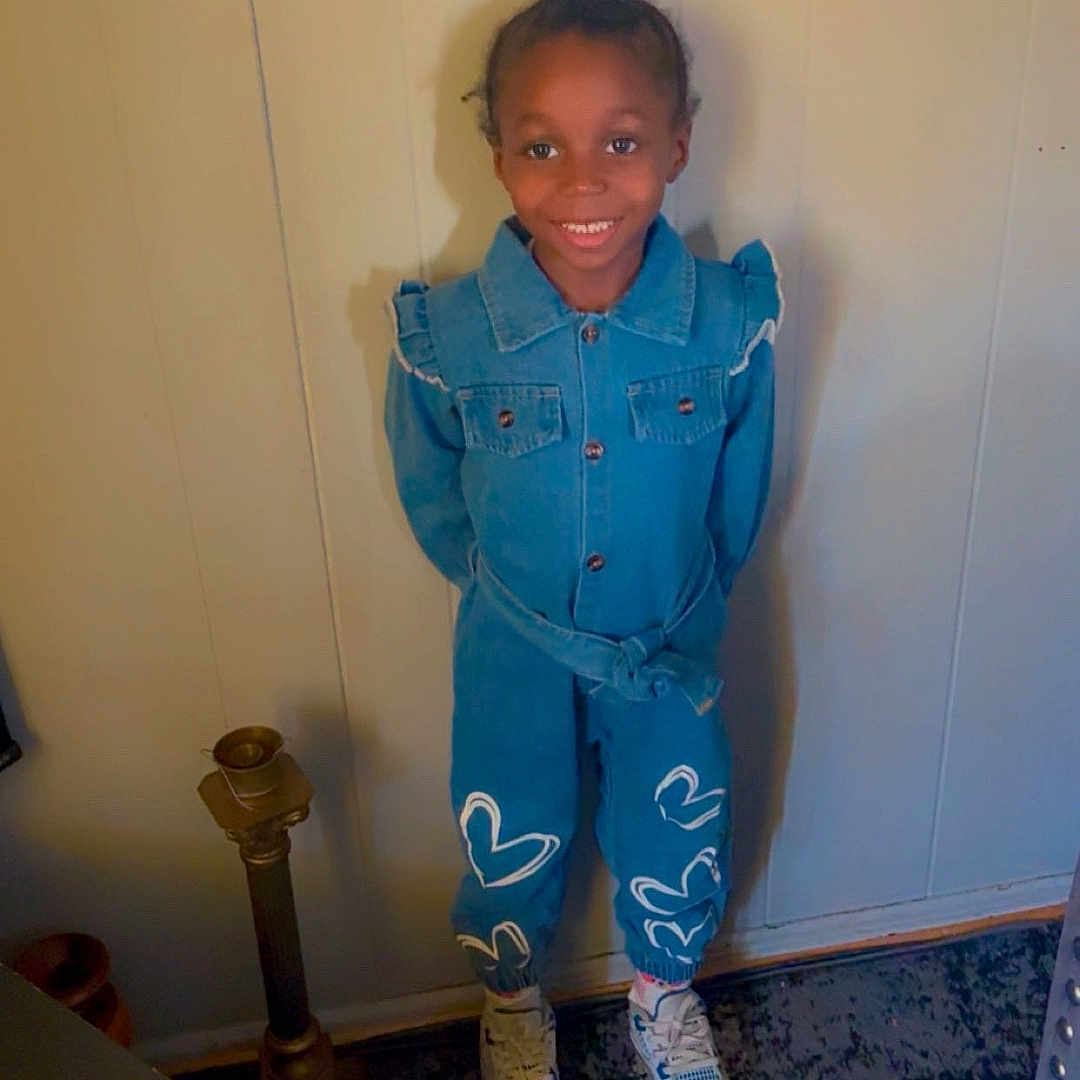 Grace is registered to the contest to win money with this photo: carpet, casual_clothing, child, cute, denim_outfit, floor, furniture, happy, heart_design, home, indoor, lamp, person, portrait, shoe, smiling, standing, table, wall, young