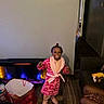 child, girl, smiling, robe, pink, slippers, living_room, electric_fireplace, gift_bag, toy, wood_floor, wall, doorway, furniture, person, happy, indoor, home, celebration, cozy