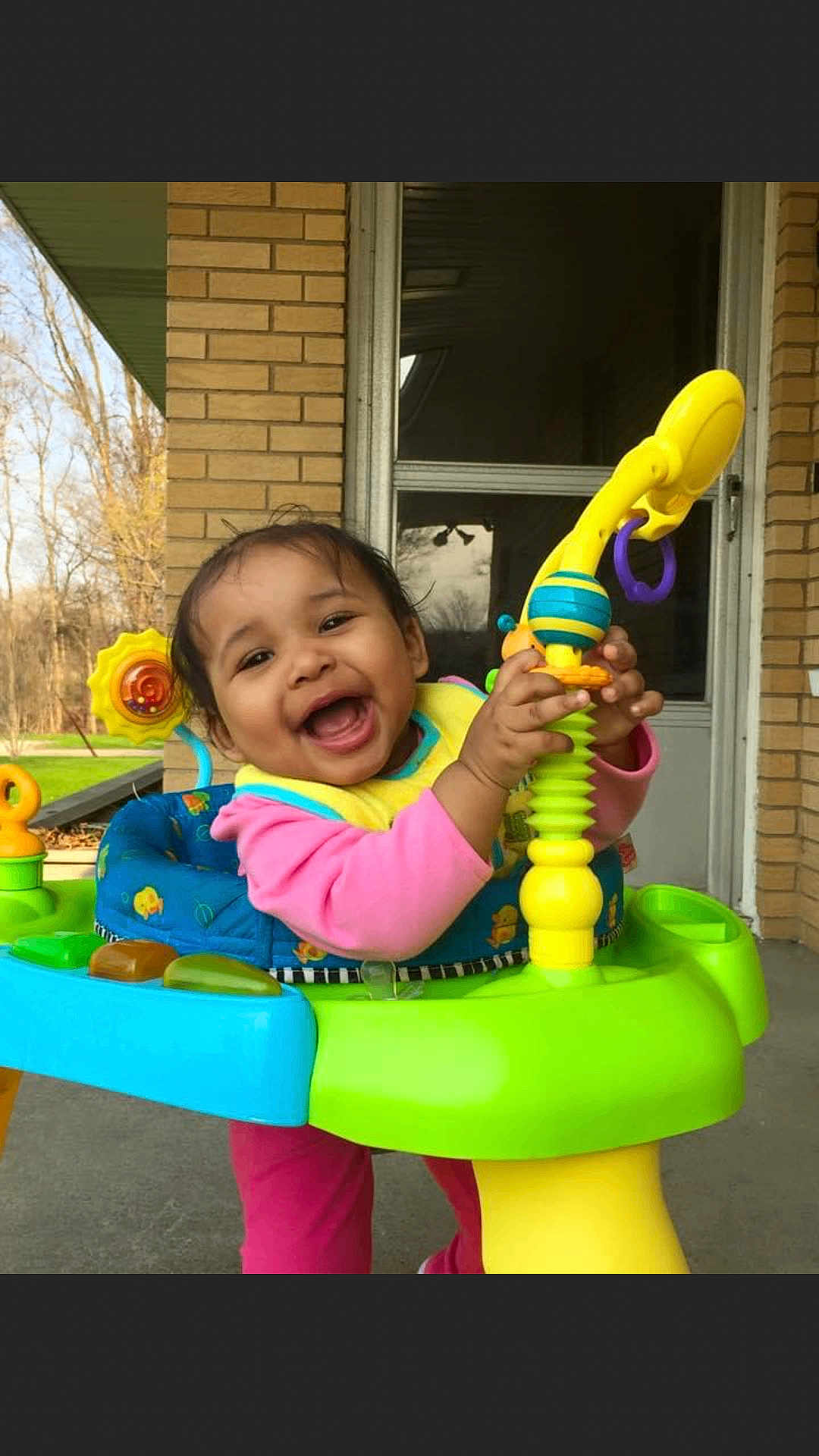 Annabelle is registered to the contest to win money with this photo: baby, child, smiling, toy, activity_center, outdoor, brick_wall, door, playtime, happy, colorful, clothing, person, infant, window, green, yellow, pink, blue, fun
